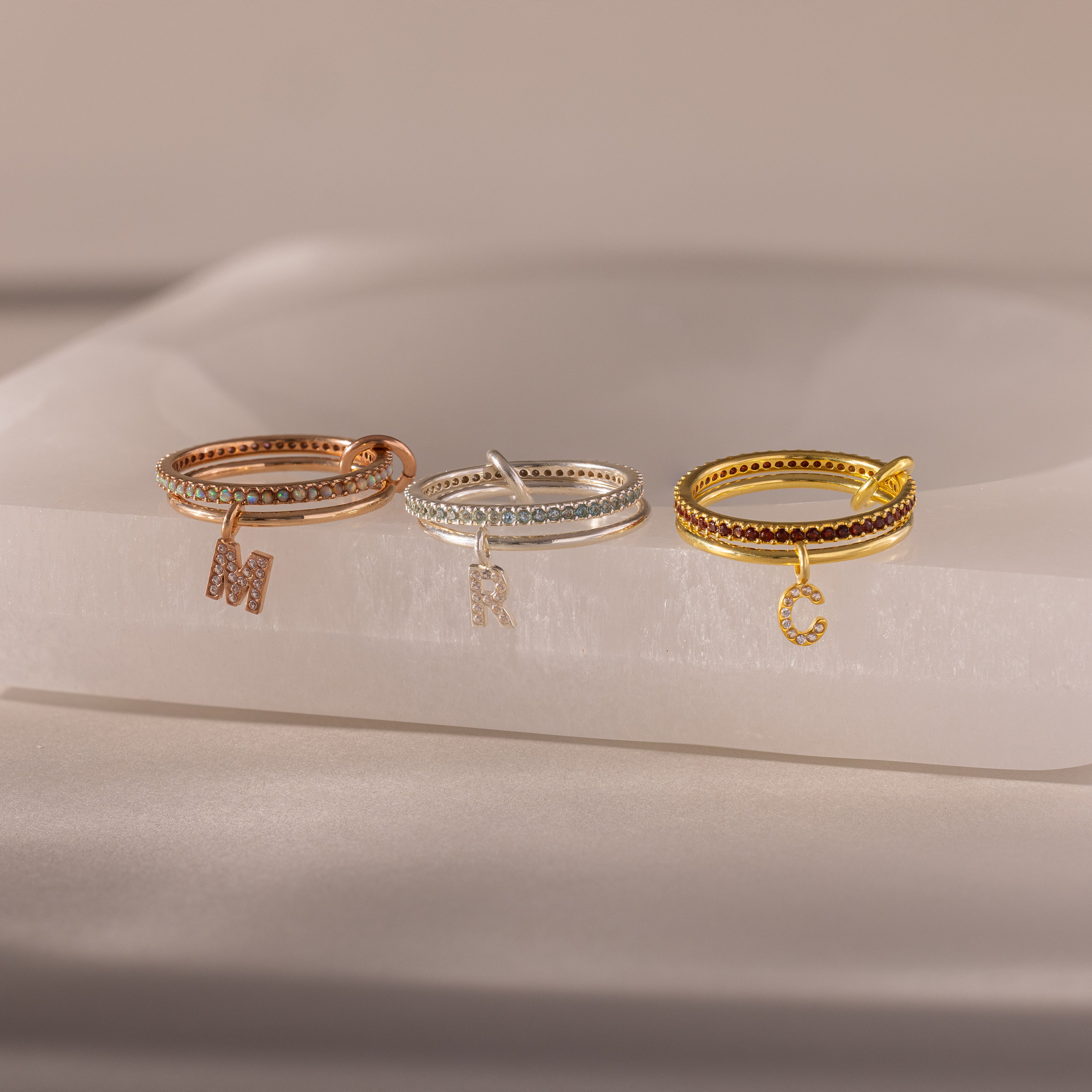 The Linked Initial Birthstone Ring Set features three rings—rose gold, silver, and gold—each with a dangling letter charm. Wear them stacked or solo for a personalized touch that adds unique style to any look.