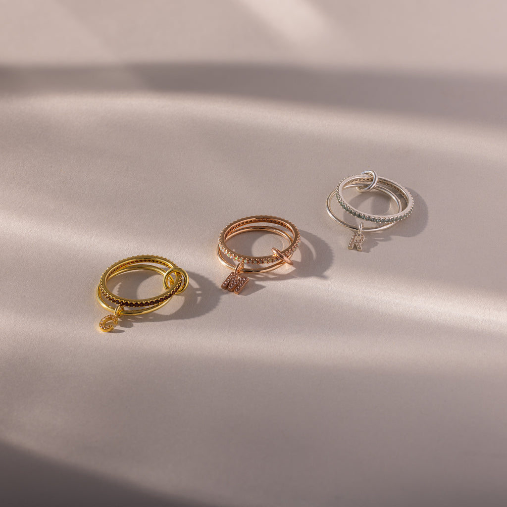 The Linked Initial Birthstone Ring Set, featuring gold, rose gold, and silver rings with personalized charms, is displayed on a softly lit beige surface to highlight its elegant birthstone beauty.