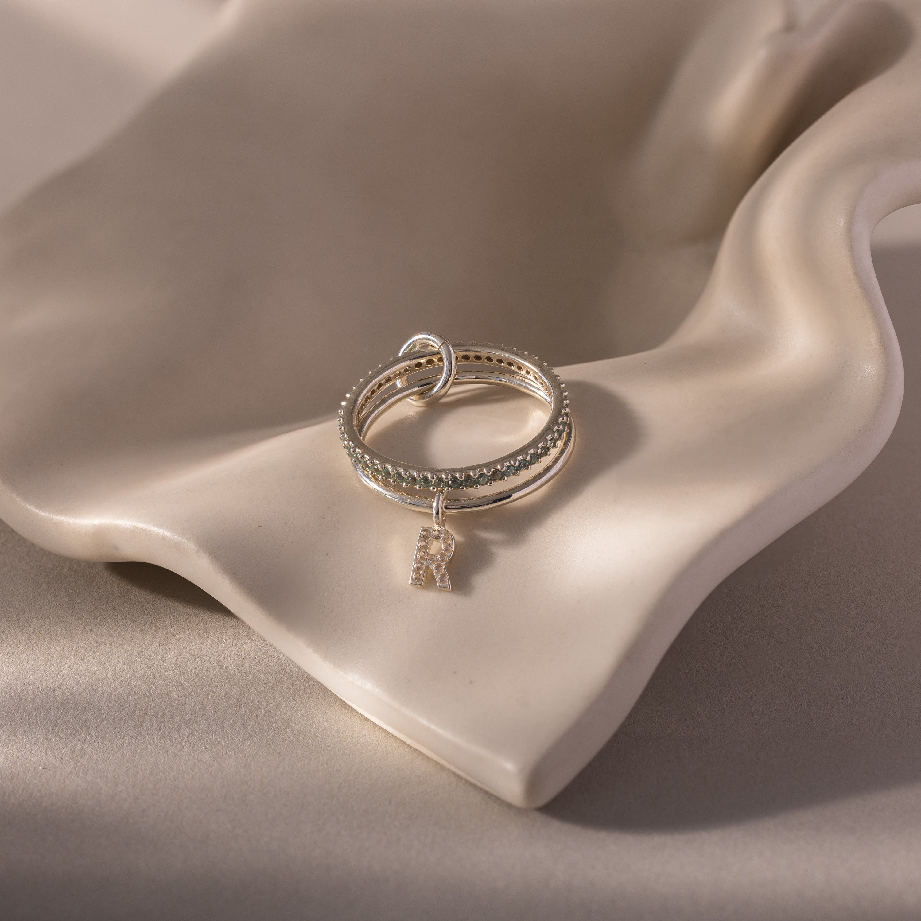 The Linked Initial Aquamarine Ring Set features silver rings with a small letter R charm, displayed on a smooth, wavy beige ceramic surface for an elegant personalized jewelry look.