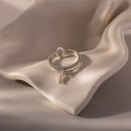 The Linked Initial Birthstone Ring Set rests on a smooth, wavy, cream-colored ceramic surface.