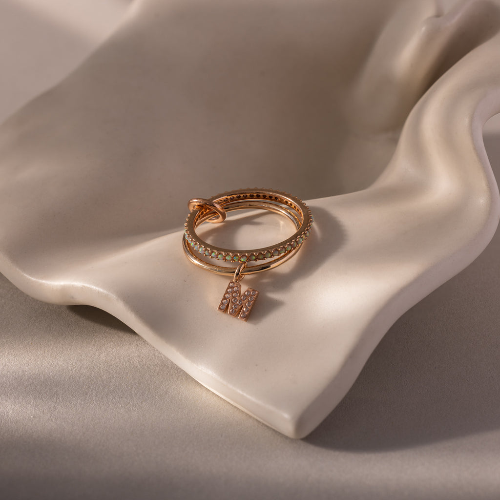 The Linked Initial Opal Ring Set features gold rings with a small M-shaped initial charm, displayed on a smooth, cream-colored ceramic surface.
