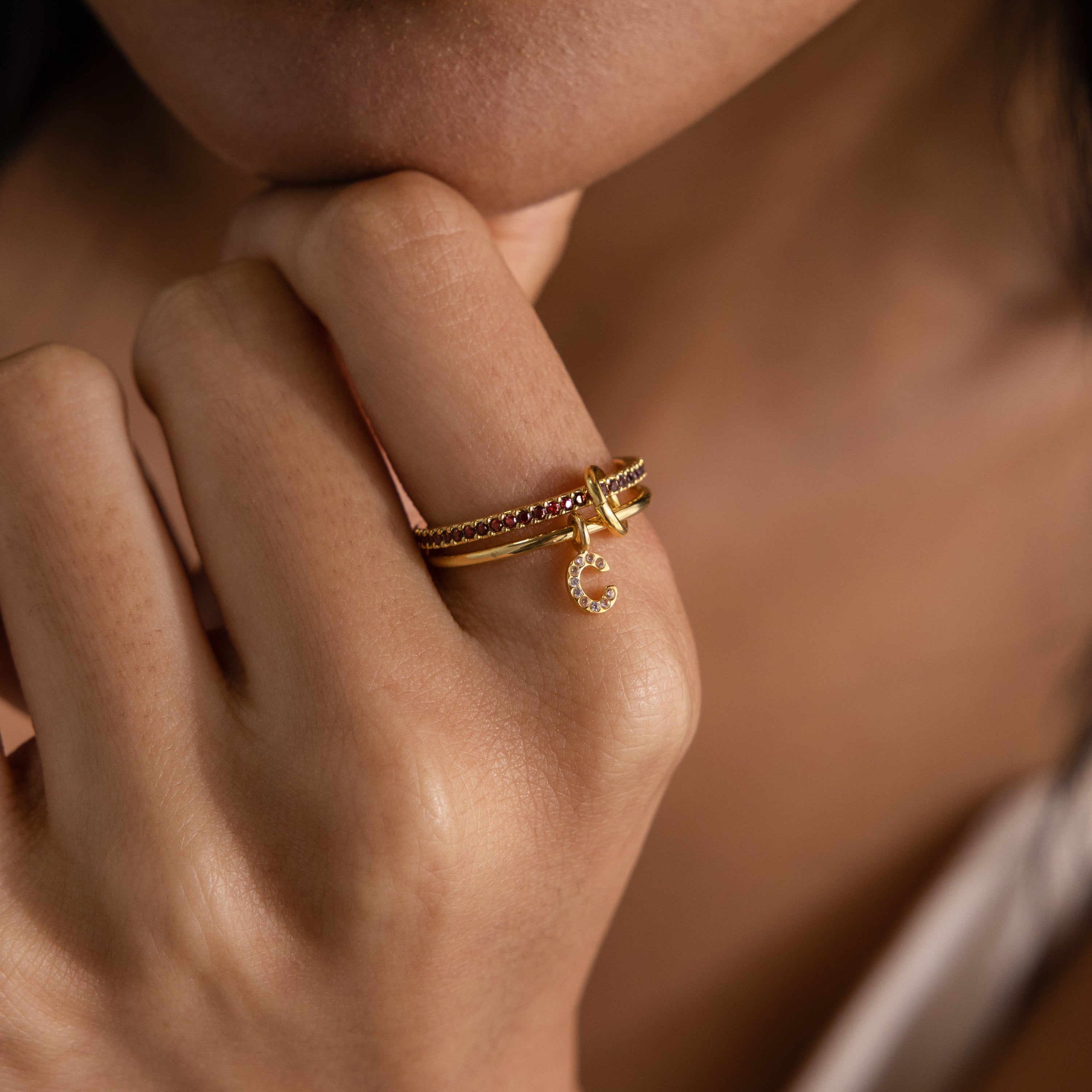 A hand wearing gold rings, including the Linked Initial Birthstone Ring Set and one with a small diamond-studded letter C charm, held near the face.