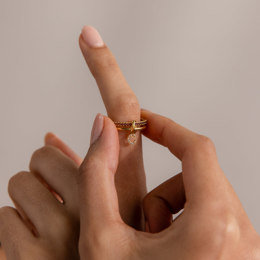 A hand slides a Linked Initial Birthstone Ring Set with a small dangling charm onto an index finger against a neutral background.