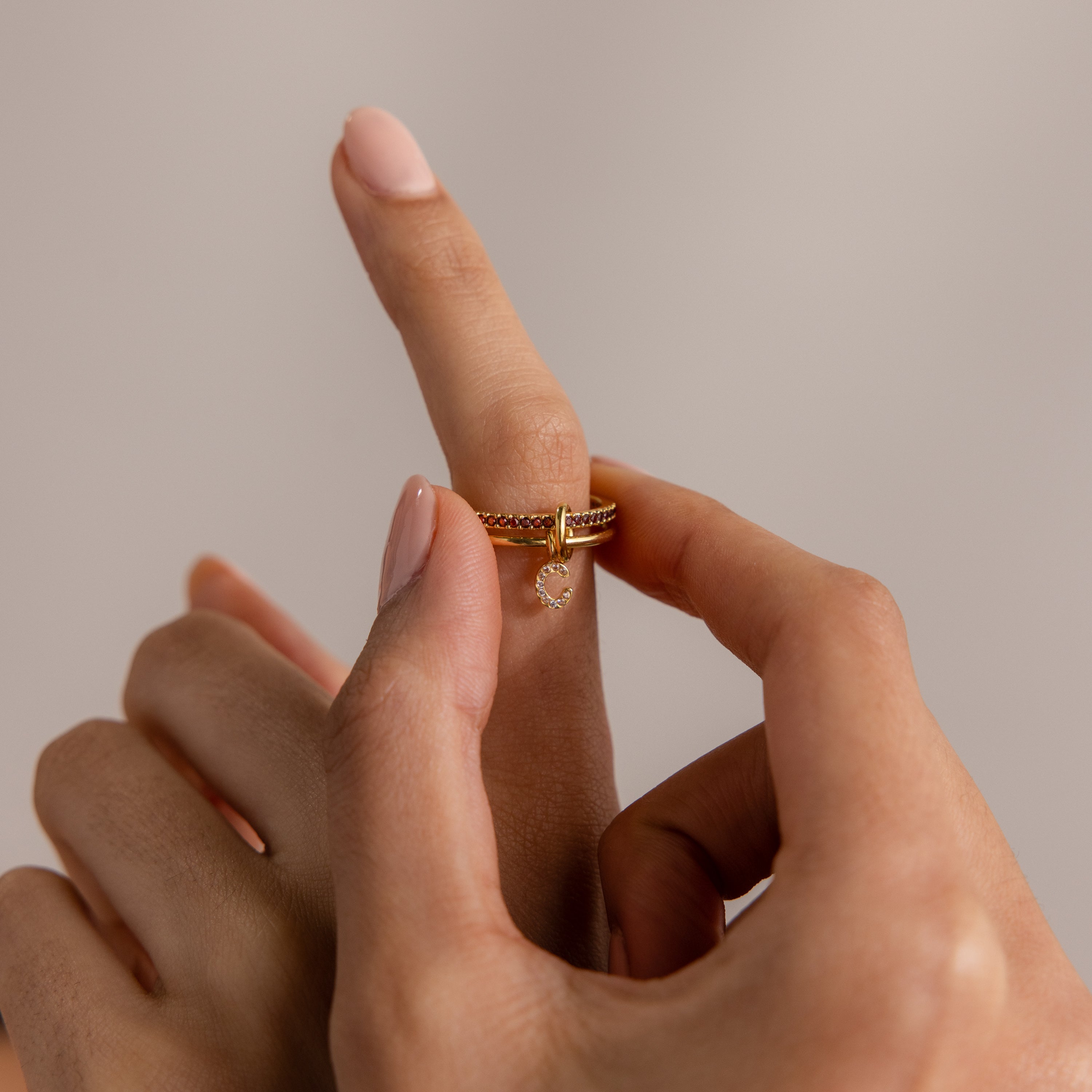 A hand slides a Linked Initial Birthstone Ring Set with a small dangling charm onto an index finger against a neutral background.