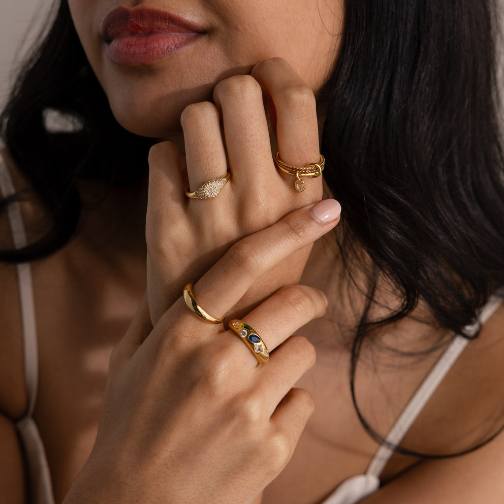 A woman with nude nail polish wears multiple gold rings, including the Linked Initial Birthstone Ring Set, and a cream-colored strap top.