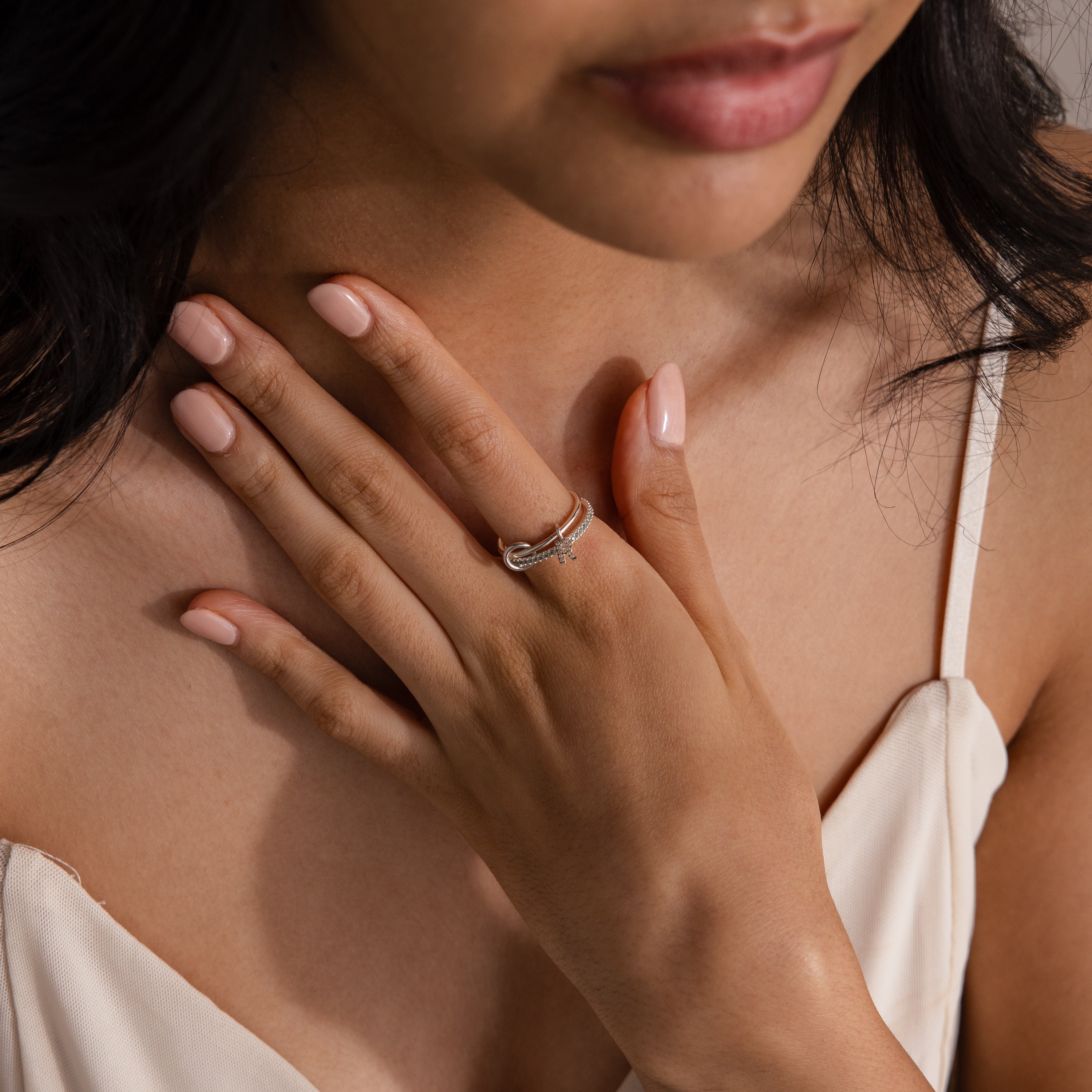 Woman in a cream dress touches her chest, displaying the Linked Initial Birthstone Ring Set—a stunning piece of personalized jewelry featuring an initial and birthstone.