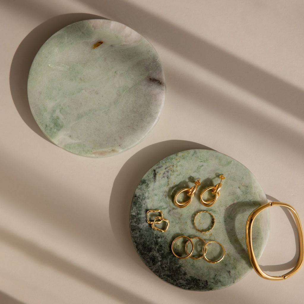 Two green Marble Jewelry Dishes display gold earrings, rings, and a bracelet on a beige surface.