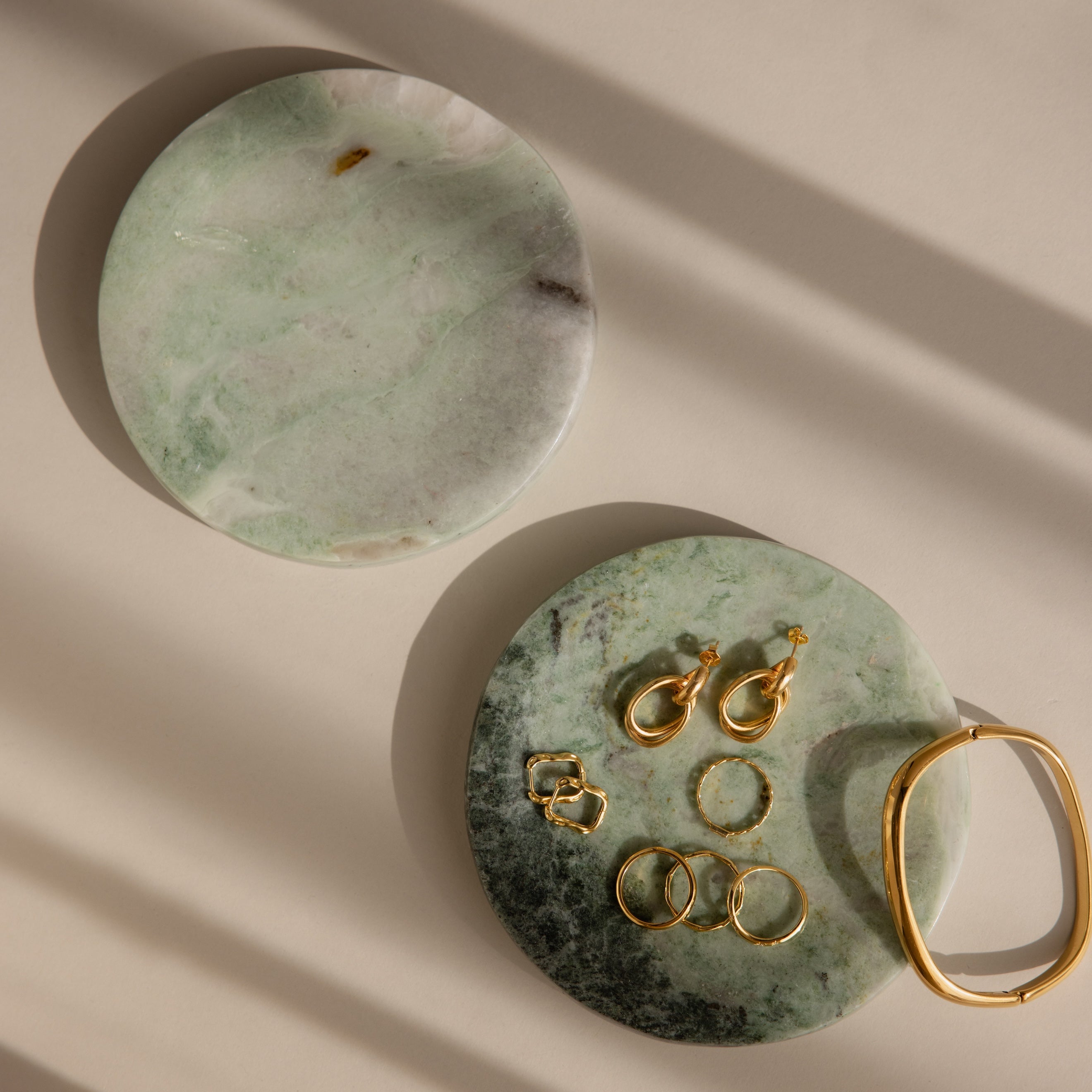 Two green Marble Jewelry Dishes display gold earrings, rings, and a bracelet on a beige surface.