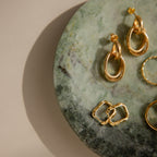 Gold earrings rest on a Green Marble Jewelry Dish, with light and shadow playing across the tray against a beige background.