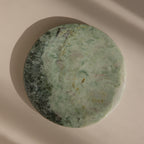 The Green Marble Jewelry Dish, featuring a round shape and greenish hues, is displayed on a beige background with soft shadows.