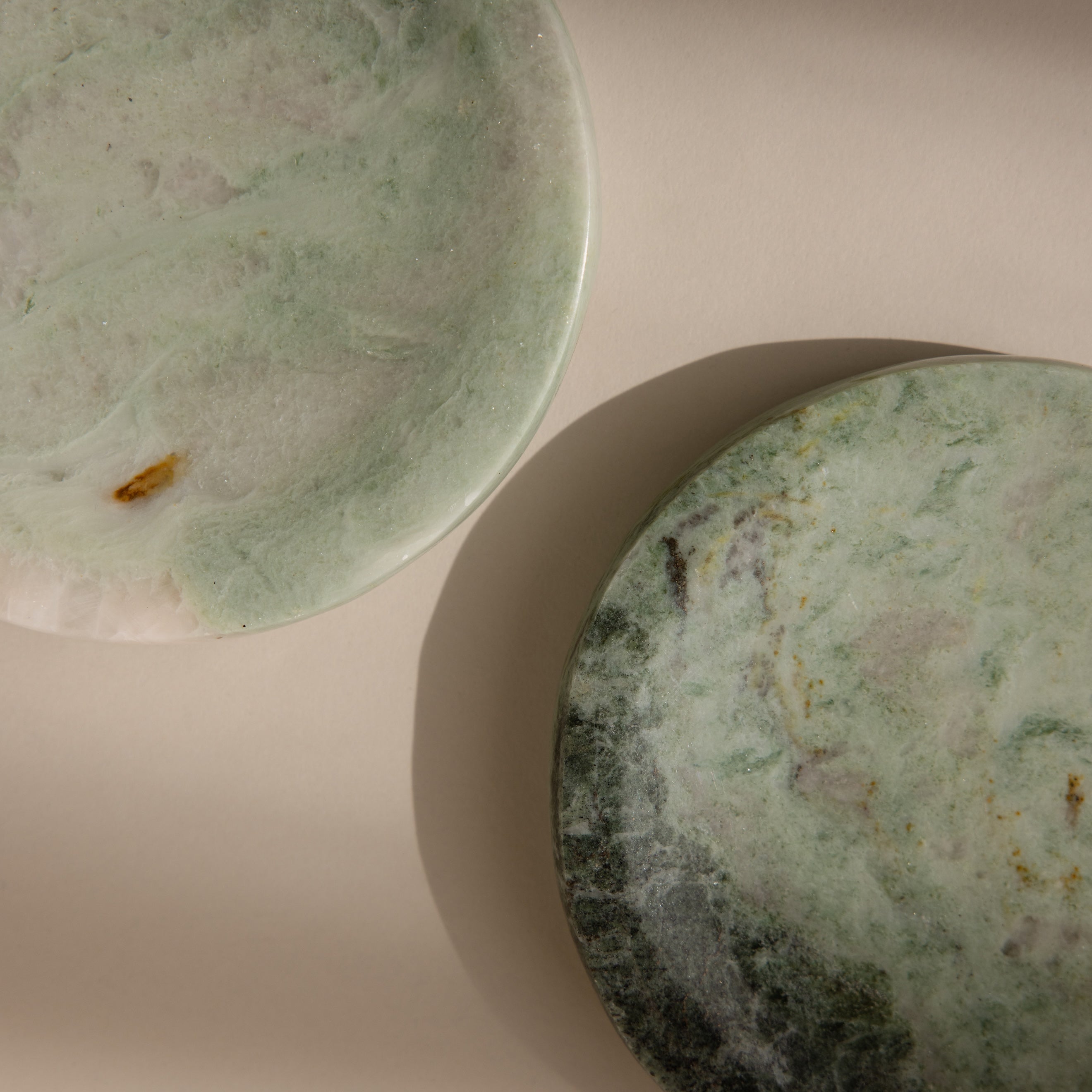The Marble Jewelry Dish, featuring two round green plates with natural patterns, serves as an elegant jewelry holder. Displayed on a beige surface and softly lit by sunlight, it adds a touch of sophistication to any space.