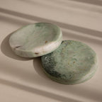 The Green Marble Jewelry Dish features a round, green marbled stone design and rests on a beige surface with soft shadows—ideal as an elegant holder for your favorite jewelry pieces.
