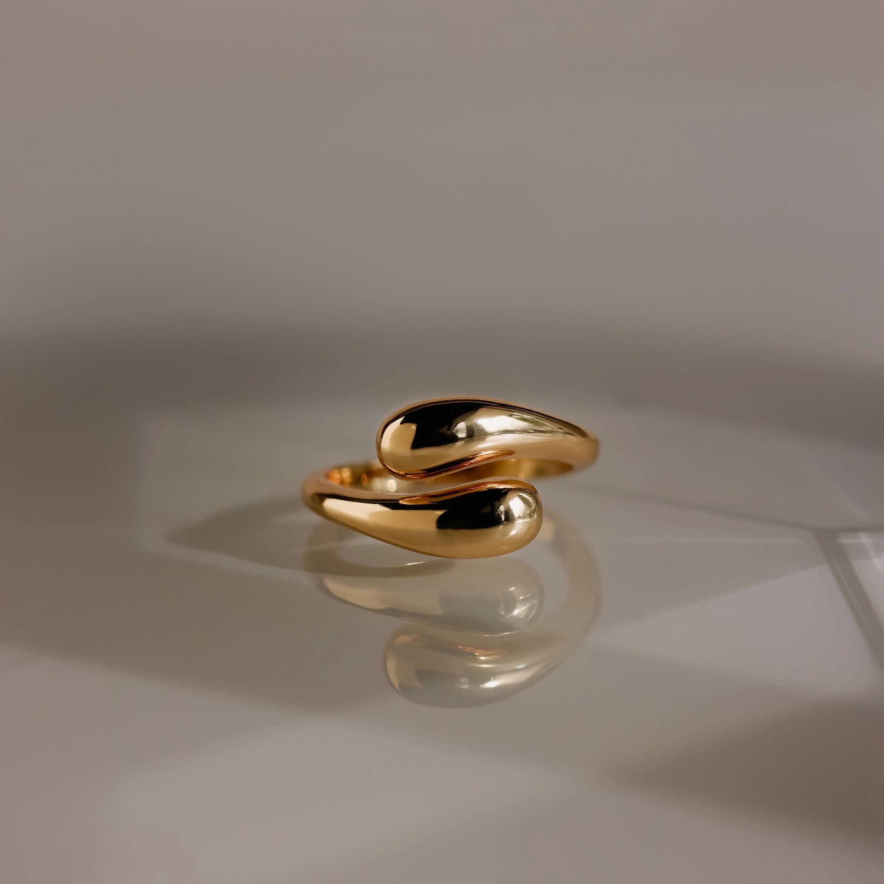 The gold Duo Dome Ring, featuring an abstract shape with curved ends and an adjustable band, is shown on a reflective surface, capturing the essence of modern sculptural jewelry.