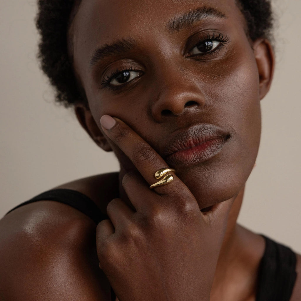 A woman with short hair touches her face and gazes thoughtfully at the camera while wearing the Duo Dome Ring, featuring an adjustable band that embodies the elegance of sculptural jewelry.