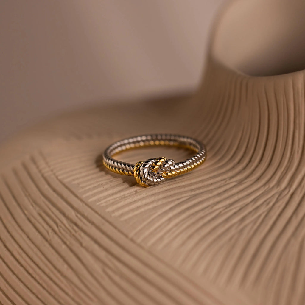 The Mixed Metal Knot Ring, ideal as a relationship gift, features gold and silver twists and rests elegantly on a textured beige surface.