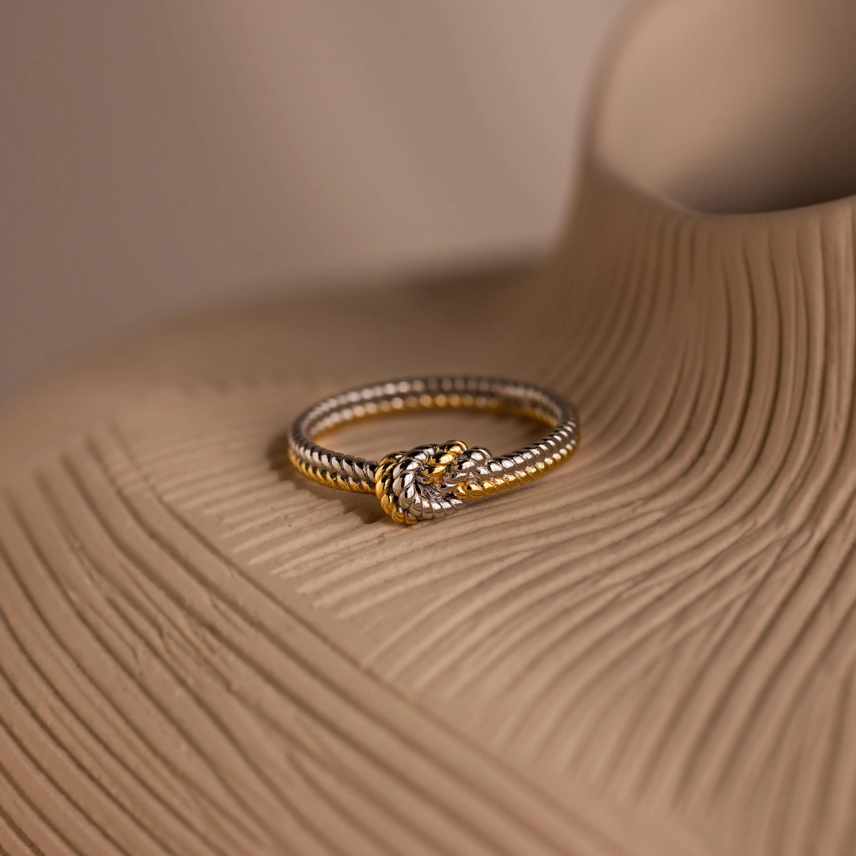 The Mixed Metal Knot Ring, ideal as a relationship gift, features gold and silver twists and rests elegantly on a textured beige surface.