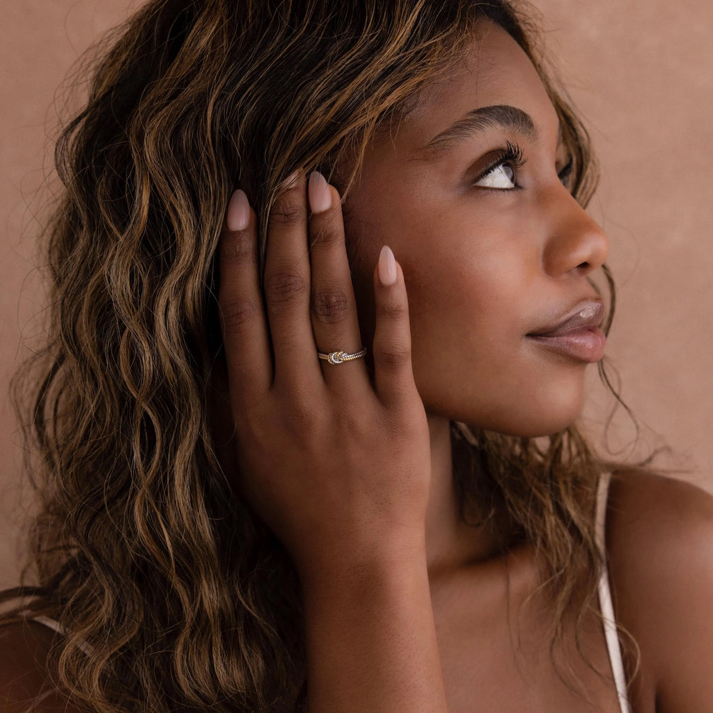 A woman with wavy hair in a beige top looks up, showing off the Mixed Metal Knot Ring in gold and silver on her left hand—a perfect relationship gift.