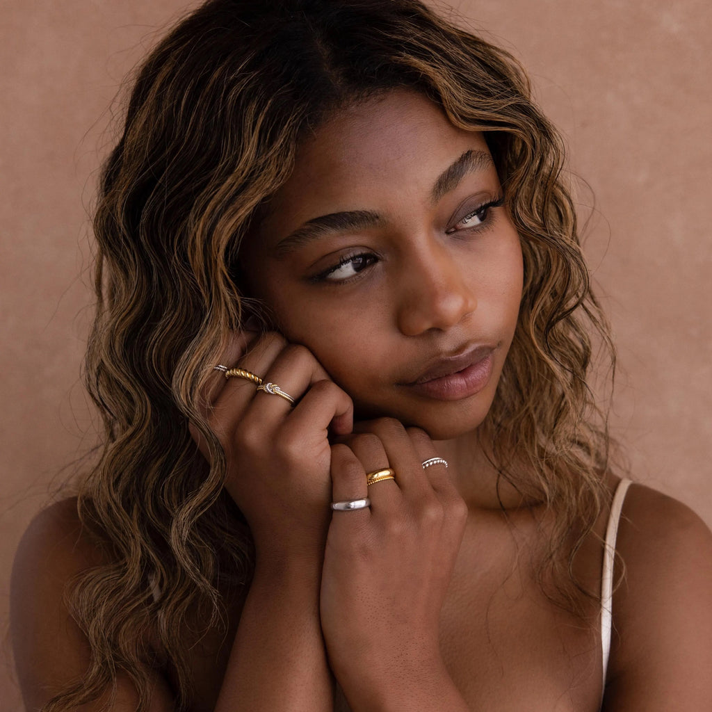 A woman with wavy hair wears the Mixed Metal Knot Ring on her finger and gazes thoughtfully to the side against a beige background—an ideal gift for celebrating relationships.