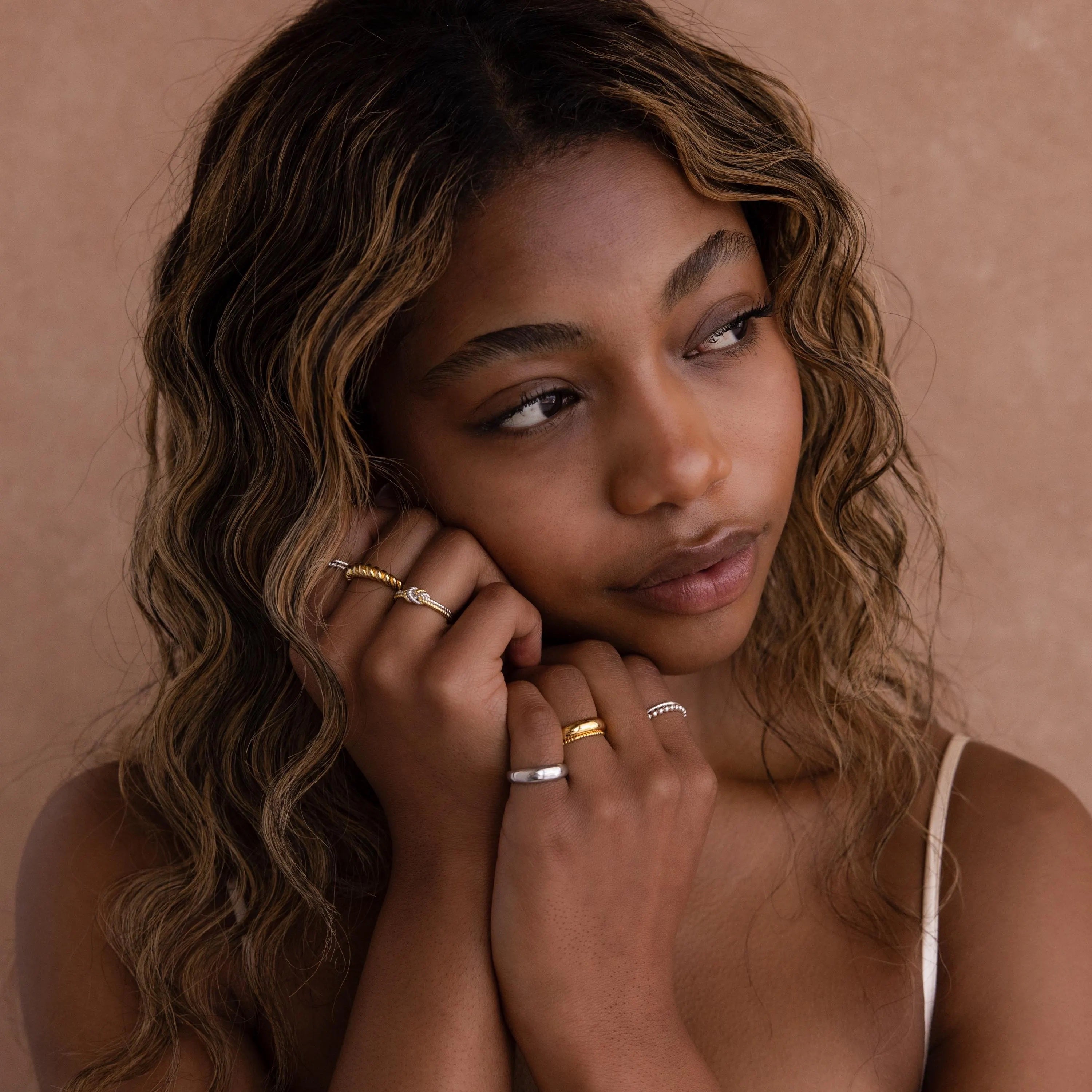 A woman with wavy hair wears the Mixed Metal Knot Ring on her finger and gazes thoughtfully to the side against a beige background—an ideal gift for celebrating relationships.