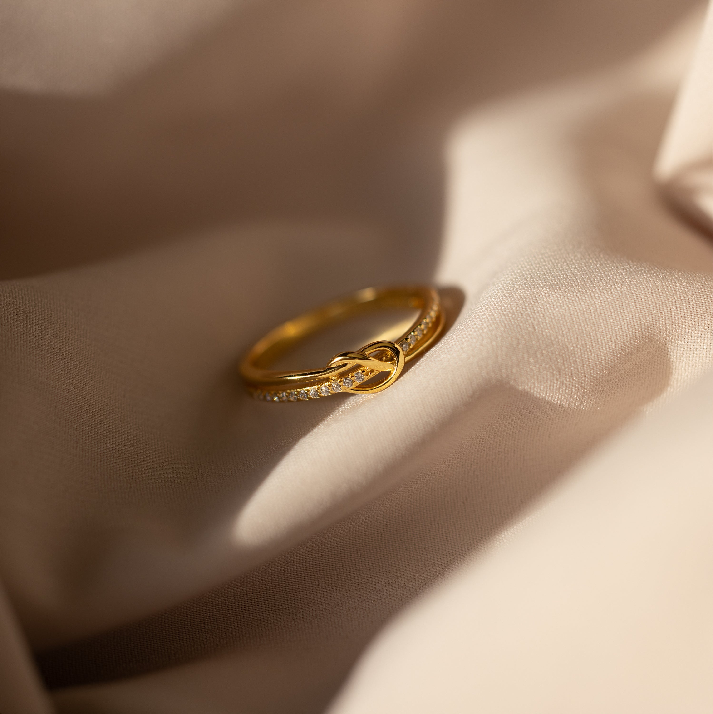 The Pave Knot Ring features an elegant knot design with sparkling accents—ideal as a jewelry gift—shown here resting on soft beige fabric in natural light.