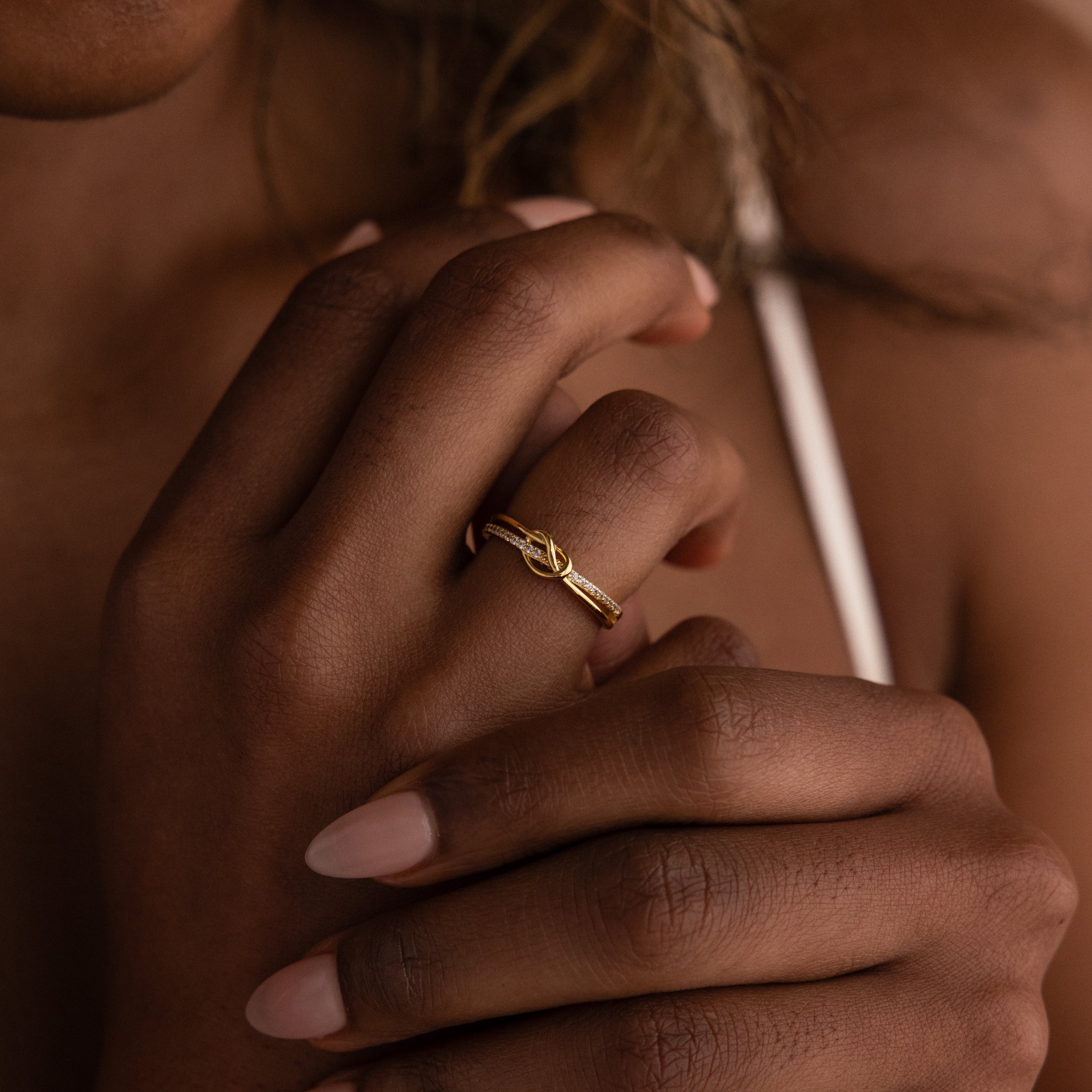 A manicured hand wears the Pave Knot Ring close to the chest—a gold ring featuring an elegant, timeless knot design and sparkling pavé, perfect as a jewelry gift.