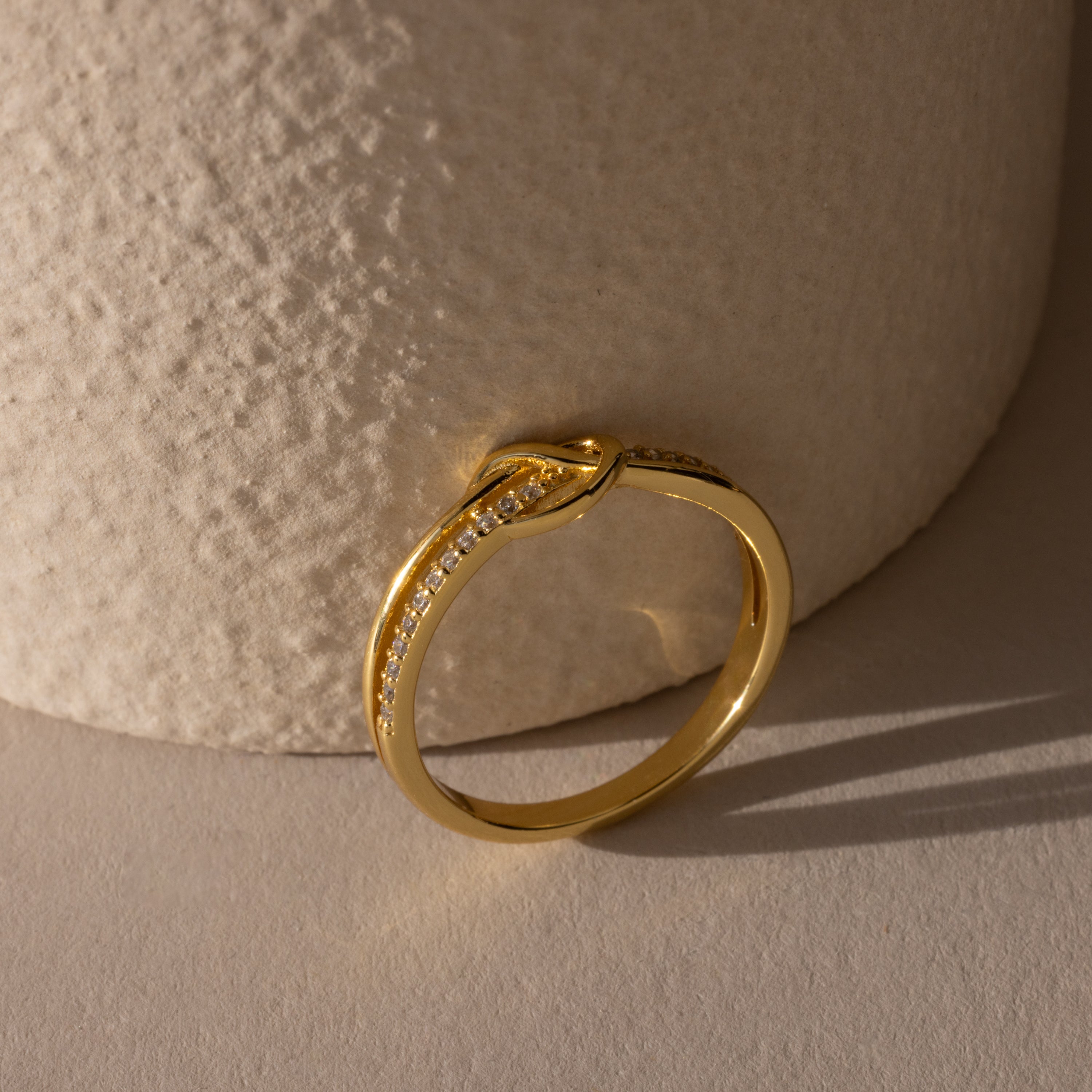 The Pave Knot Ring, featuring small embedded diamonds and a striking knot design, rests on a textured beige surface—an ideal jewelry gift.