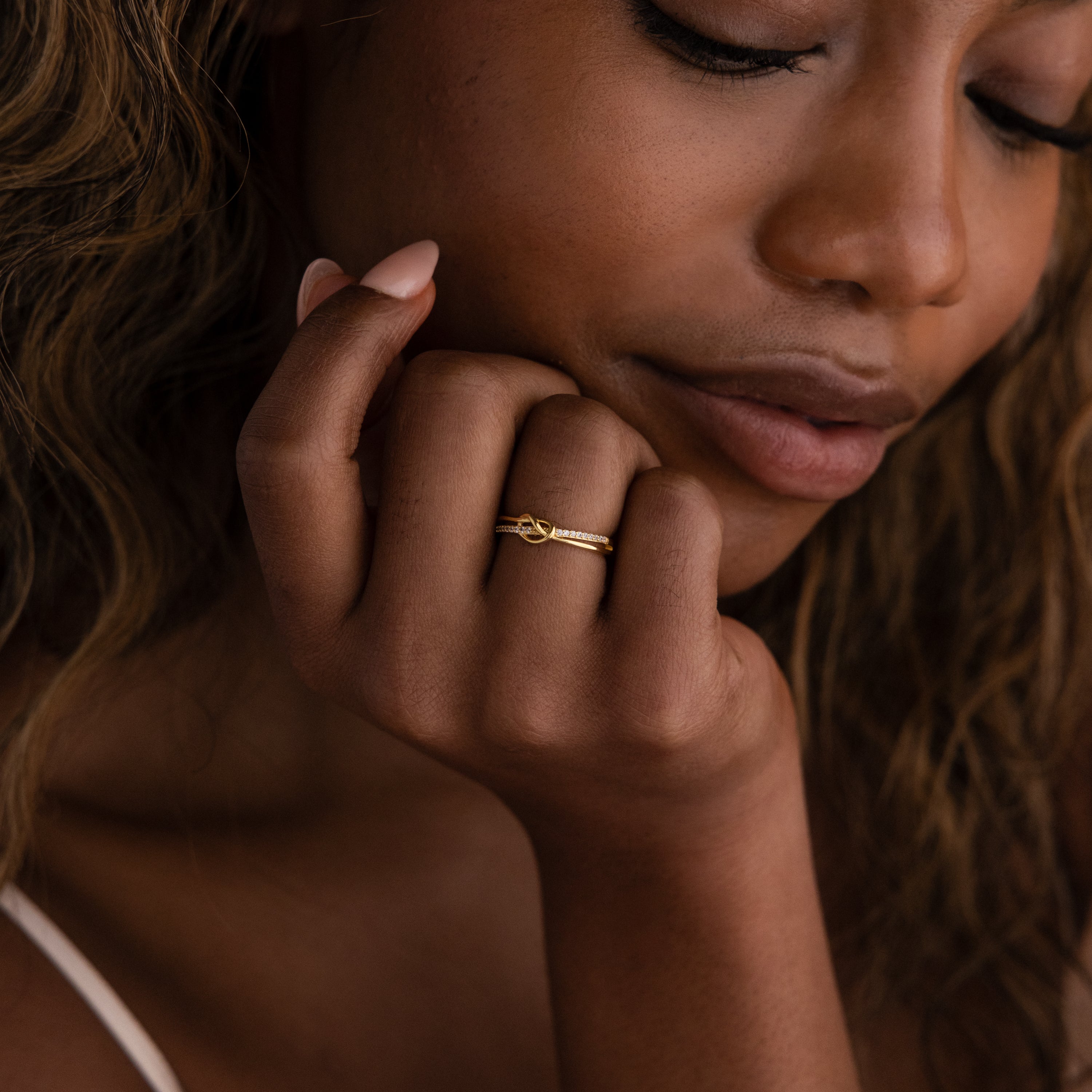 A woman with long hair gently touches her face and gazes downward, showcasing the Pave Knot Ring.
