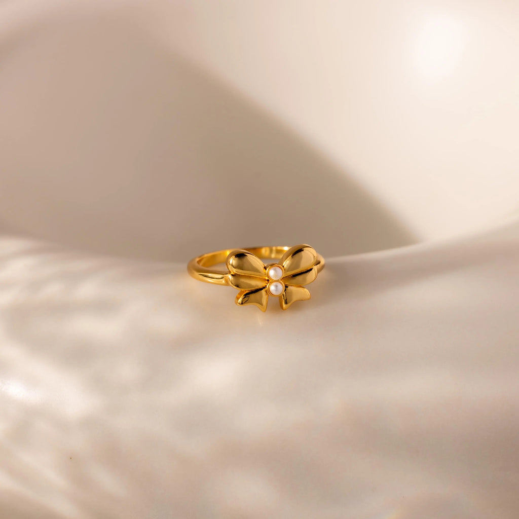 The gold Bow Pearl Ring is displayed on a softly lit, curved cream surface, highlighting its delicate design.