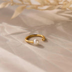 The Open Pearl & Diamond Ring, featuring a gold adjustable band with a pearl and small diamond, rests on a softly shadowed surface with dried leaves above.