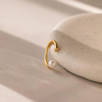 The Open Pearl & Diamond Ring features a delicate pearl and diamond, displayed on a smooth, light stone atop a beige surface—an elegant, adjustable ring with timeless style.