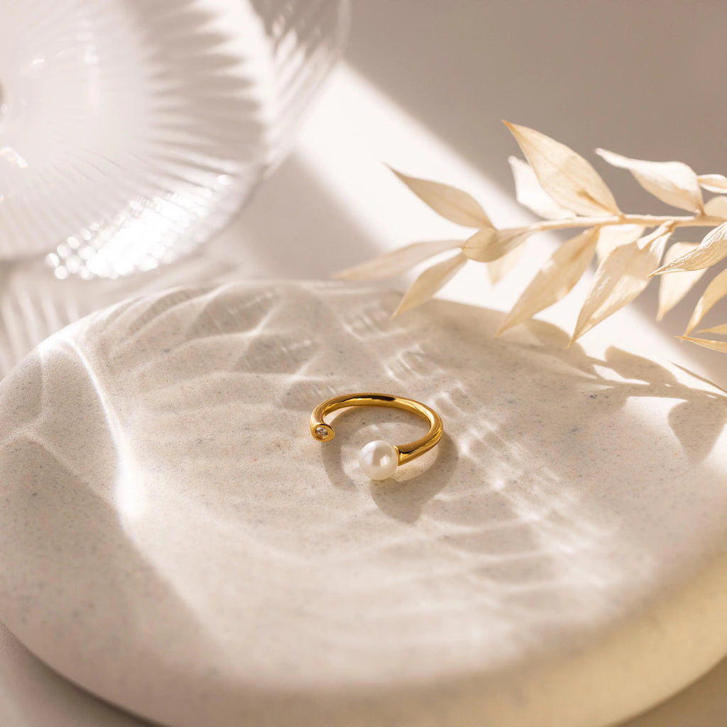 The Open Pearl & Diamond Ring, featuring an adjustable gold band with a luminous pearl, is displayed on a round stone with soft lighting and dried leaves in the background.