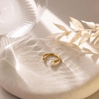 The Open Pearl & Diamond Ring, featuring an adjustable gold band with a luminous pearl, is displayed on a round stone with soft lighting and dried leaves in the background.