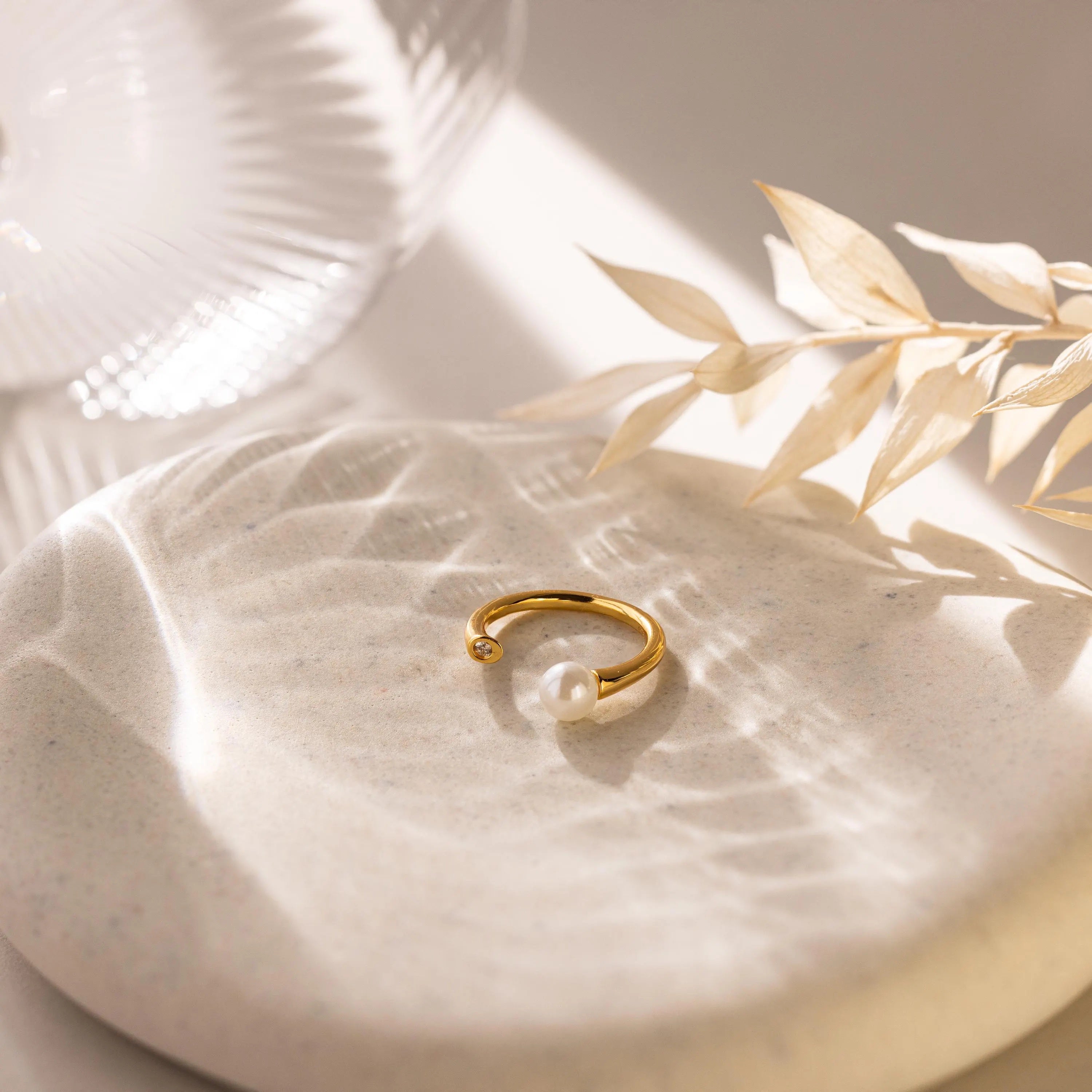 The Open Pearl & Diamond Ring, featuring an adjustable gold band with a luminous pearl, is displayed on a round stone with soft lighting and dried leaves in the background.