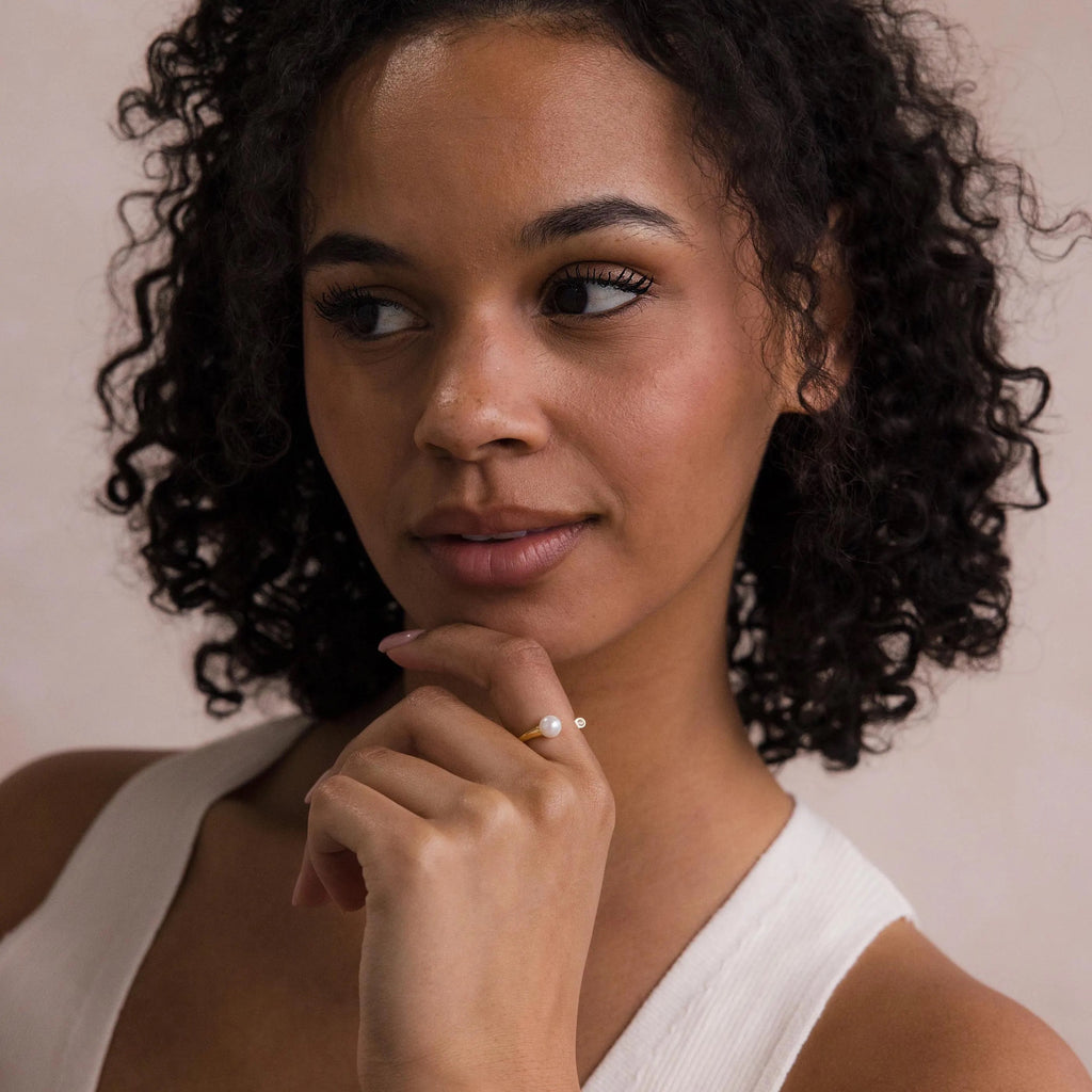 A woman with curly hair in a white top touches her chin and looks to the side, elegantly displaying the Open Pearl & Diamond Ring.