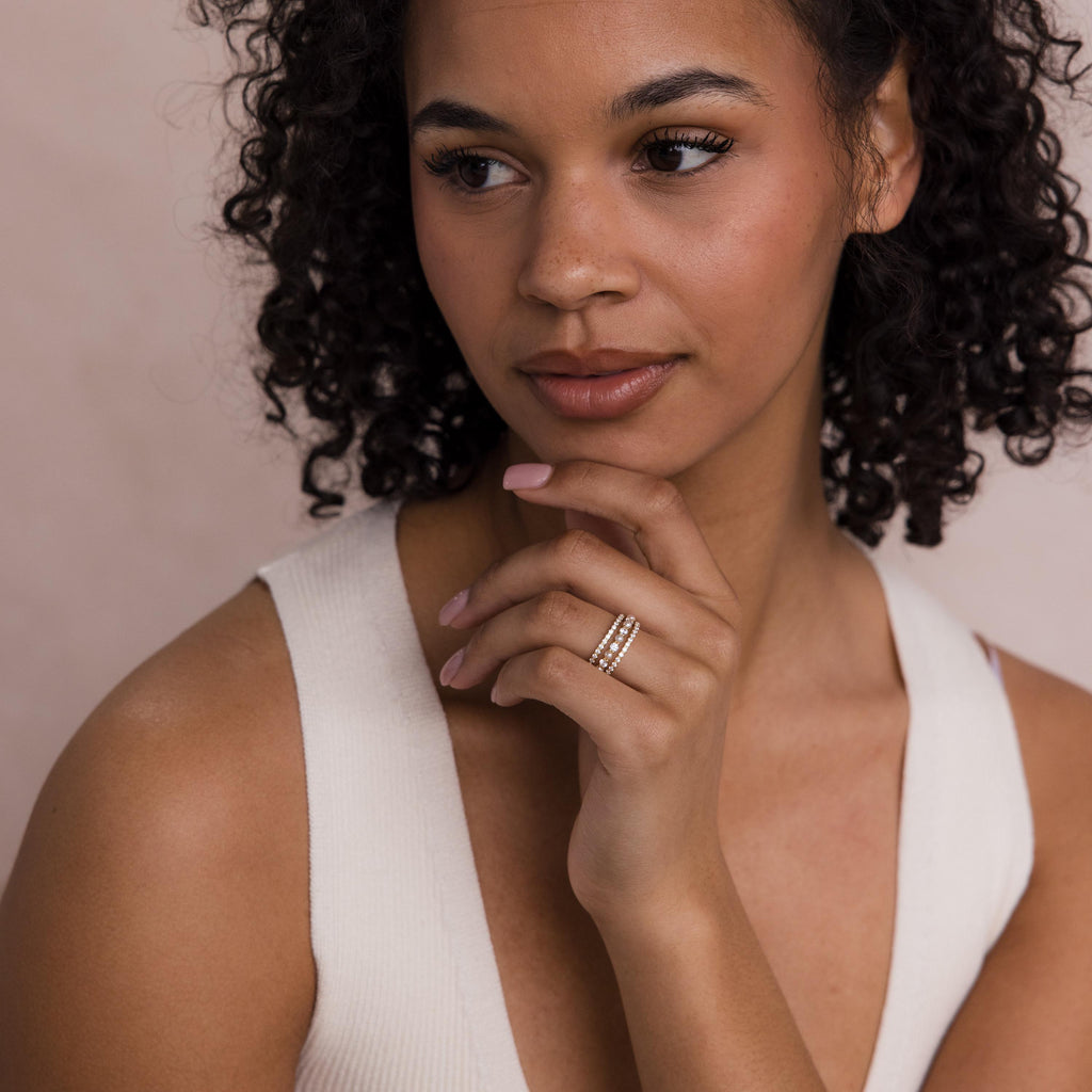 A woman with curly hair in a white top touches her chin, elegantly showcasing the Diamond Eternity Ring Set on her finger.