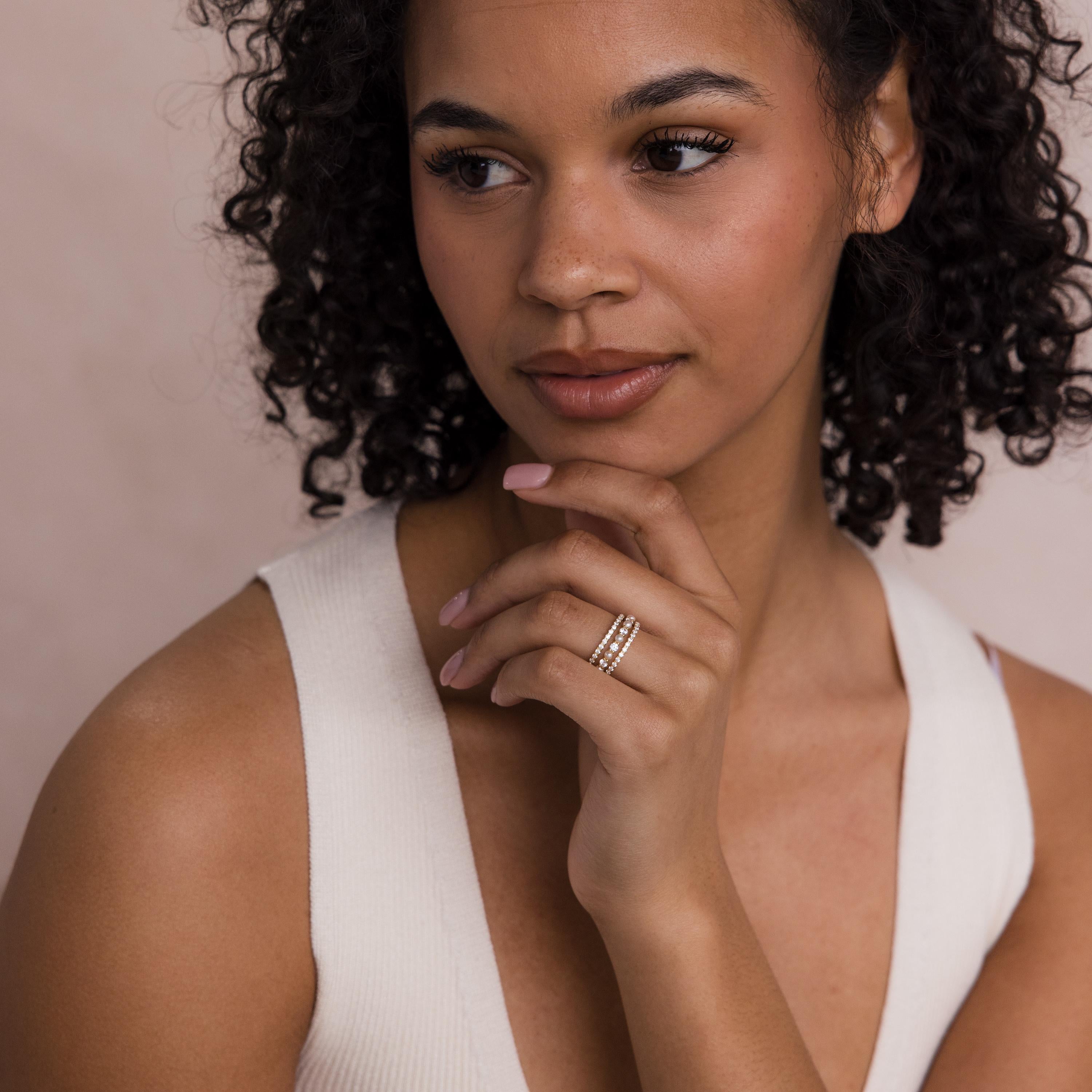 A woman with curly hair in a white top touches her chin, elegantly showcasing the Diamond Eternity Ring Set on her finger.