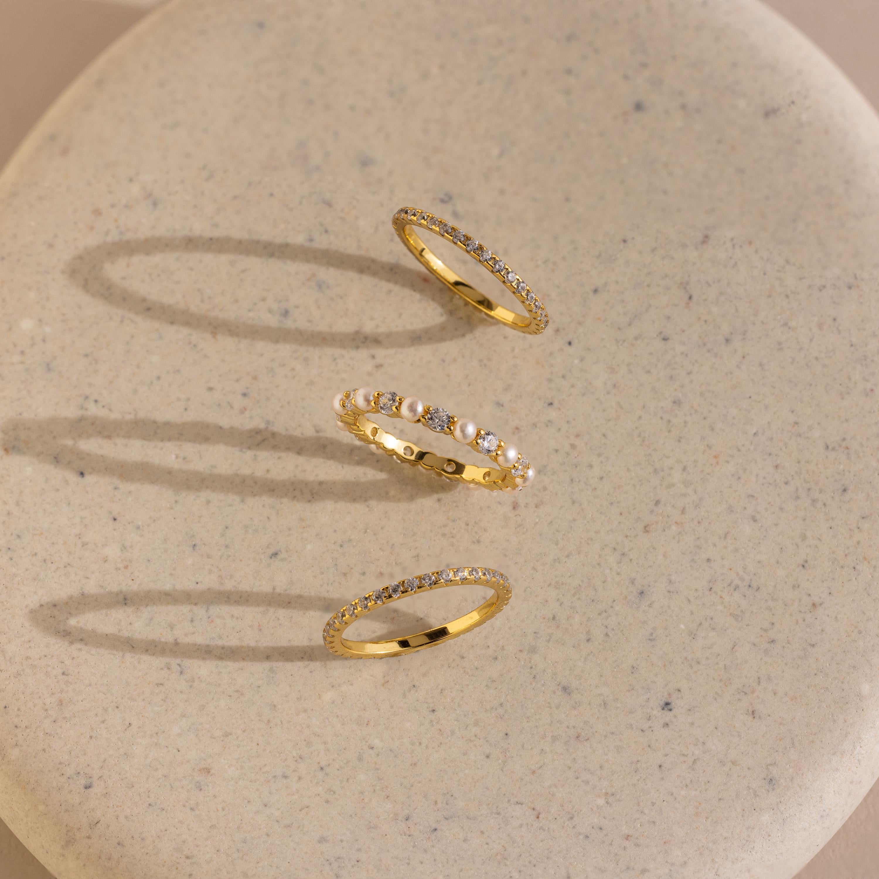 The Diamond Eternity Ring Set, featuring two gold diamond bands and a pearl ring, is elegantly displayed on a beige speckled surface.