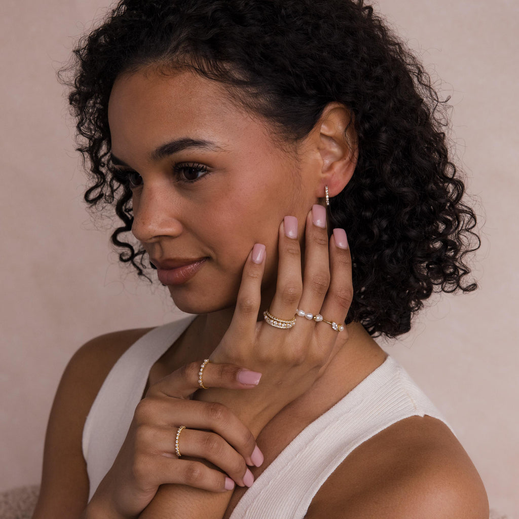 A woman with curly hair poses with her hands near her face, wearing gold rings, earrings, and the Diamond Eternity Ring Set.