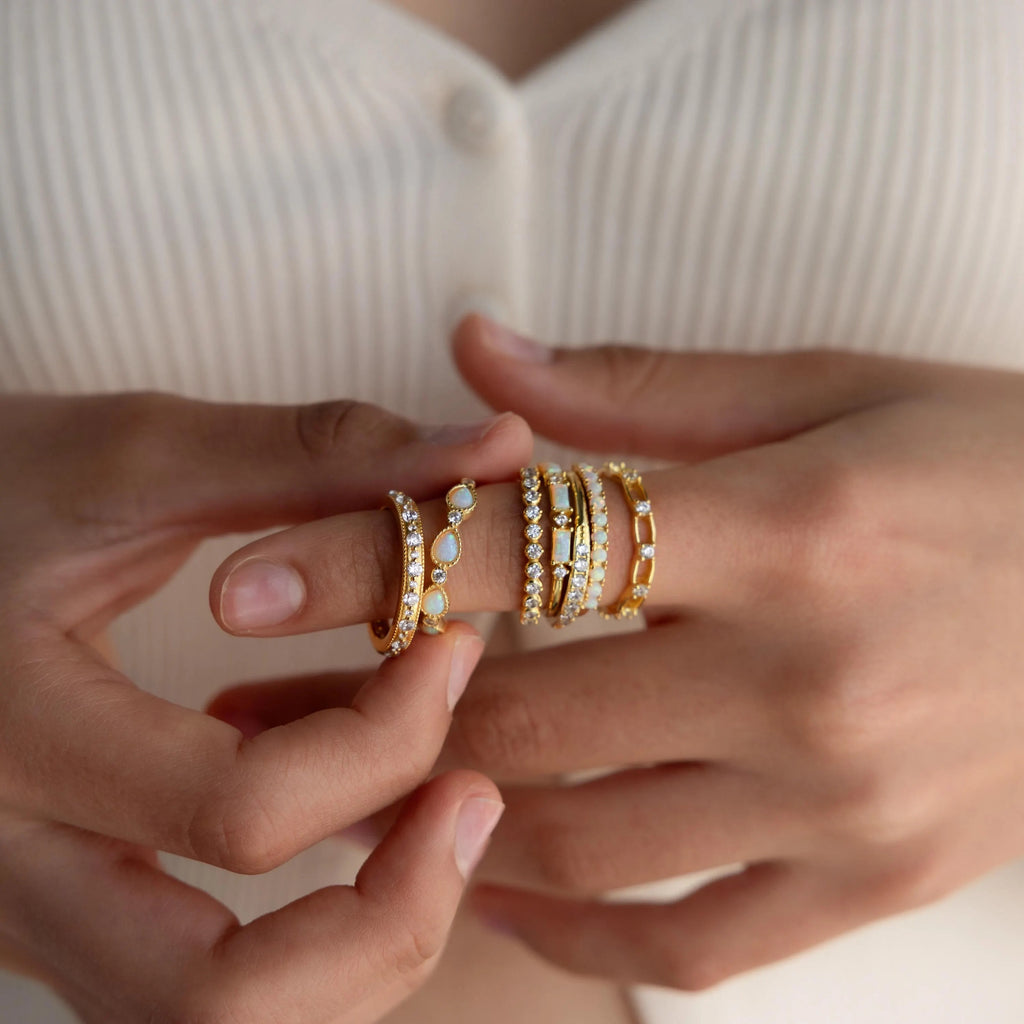 A person with several gold and gemstone rings, including the Opal Diamond Teardrop Ring, on their fingers holds their hands close to their chest.