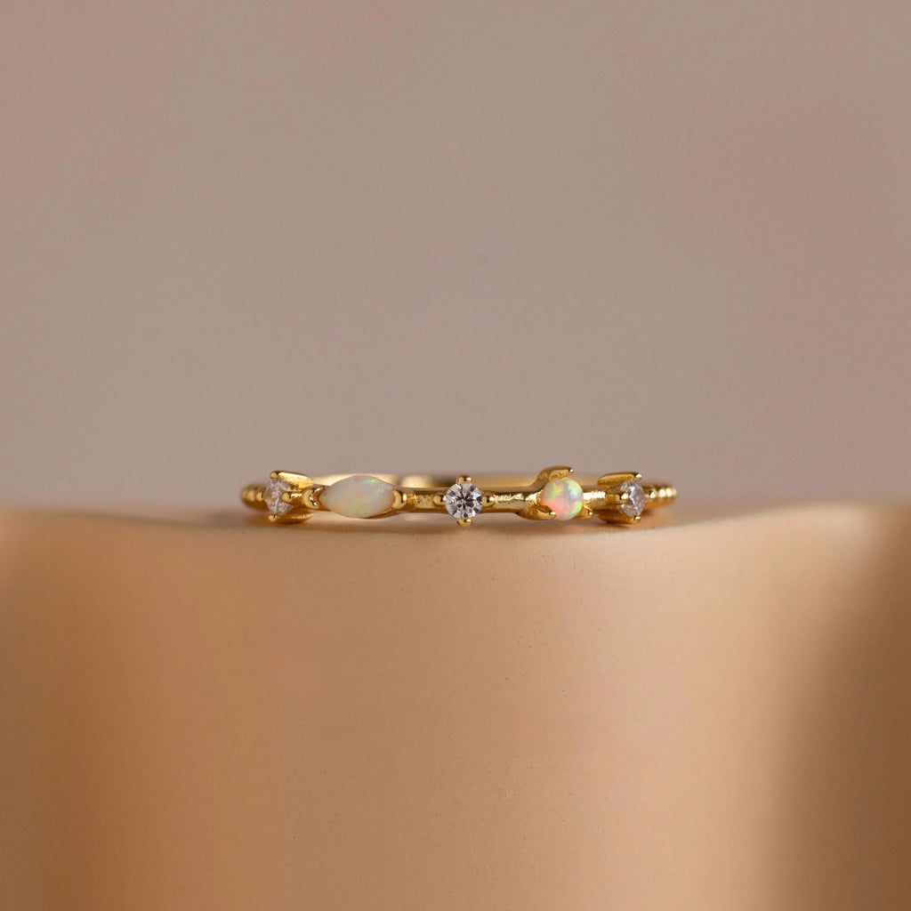 The Mixed Opal & Diamond Ring features a gold band set with small opals and diamond stones, shown on a beige surface against a neutral background.