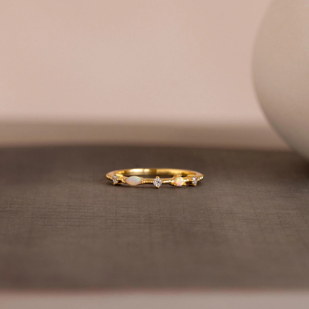 The Mixed Opal & Diamond Ring, a gold band set with opals and diamonds, is displayed on a textured gray surface against a softly blurred background.