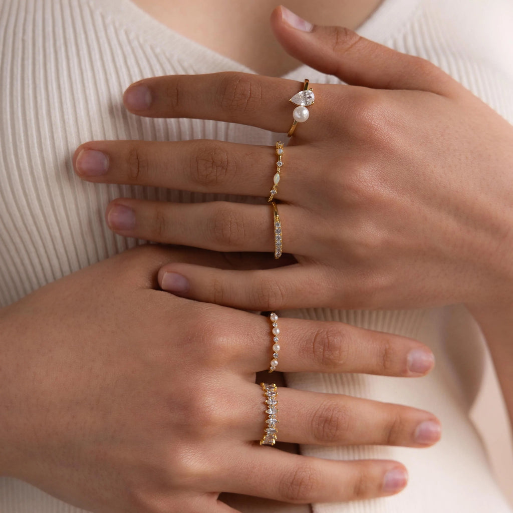Close-up of hands adorned with several gold rings, featuring the Mixed Opal & Diamond Ring set with pearls, gemstones, and opals against a cream-colored top.