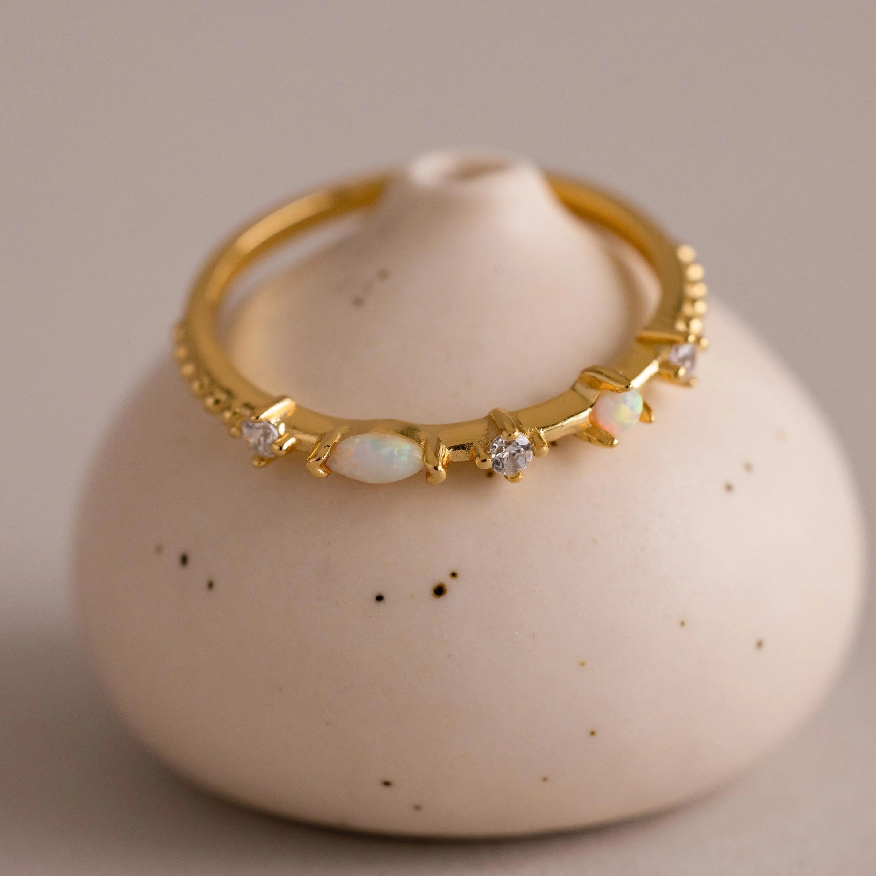 The Mixed Opal & Diamond Ring features a gold band set with opals and diamond-like stones, elegantly displayed on a round white ceramic holder.