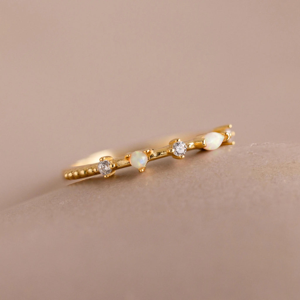 The Mixed Opal & Diamond Ring showcases a gold band with small opals and diamonds in a delicate, minimalist design against a neutral backdrop.
