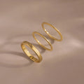 The Simple Ring Set features three thin gold stacking rings arranged in a row on a smooth beige surface with soft shadows, showcasing their elegant minimalist style.