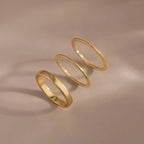 The Simple Ring Set features three thin gold stacking rings arranged in a row on a smooth beige surface with soft shadows, showcasing their elegant minimalist style.
