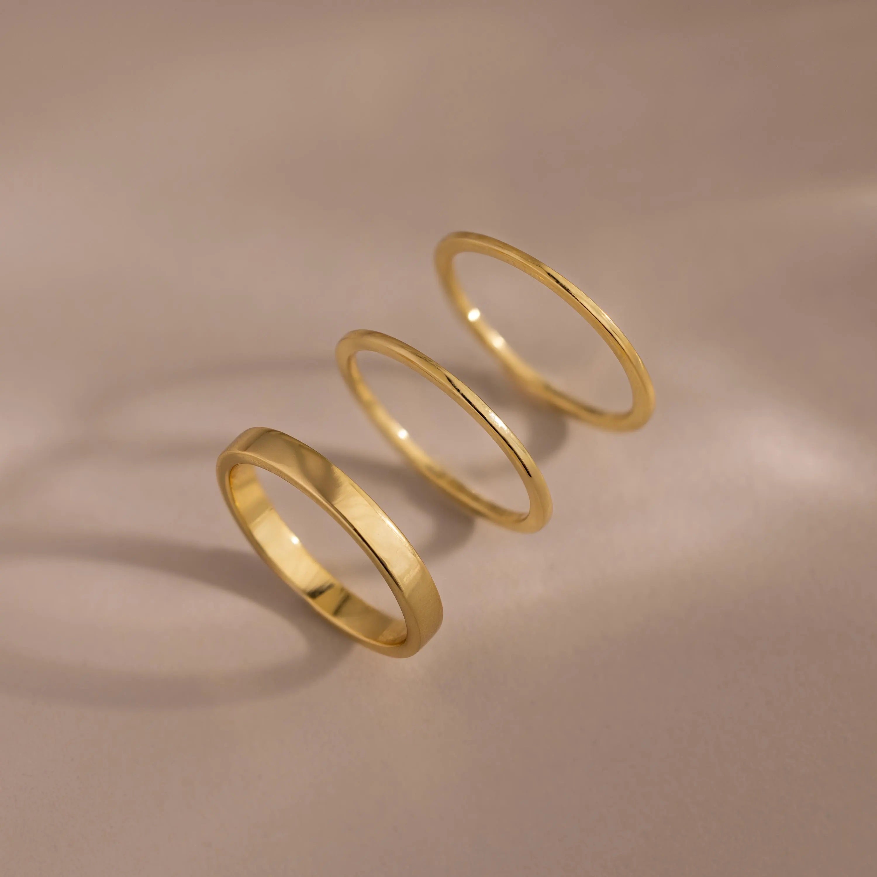 The Simple Ring Set features three thin gold stacking rings arranged in a row on a smooth beige surface with soft shadows, showcasing their elegant minimalist style.