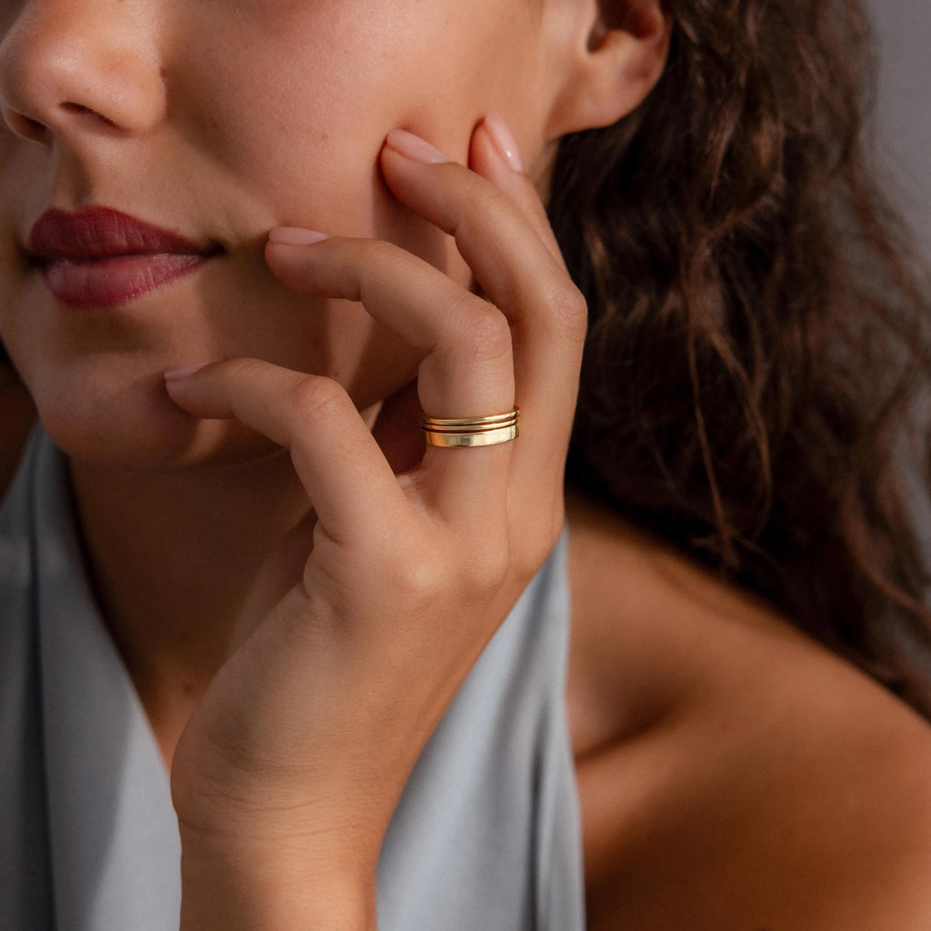 A woman with curly hair touches her face, wearing the Simple Ring Set on her finger and a light gray top.