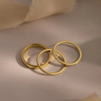 The Simple Ring Set features three elegant gold rings overlapping on a light surface with soft beige fabric in the background, creating a perfect look for stacking.