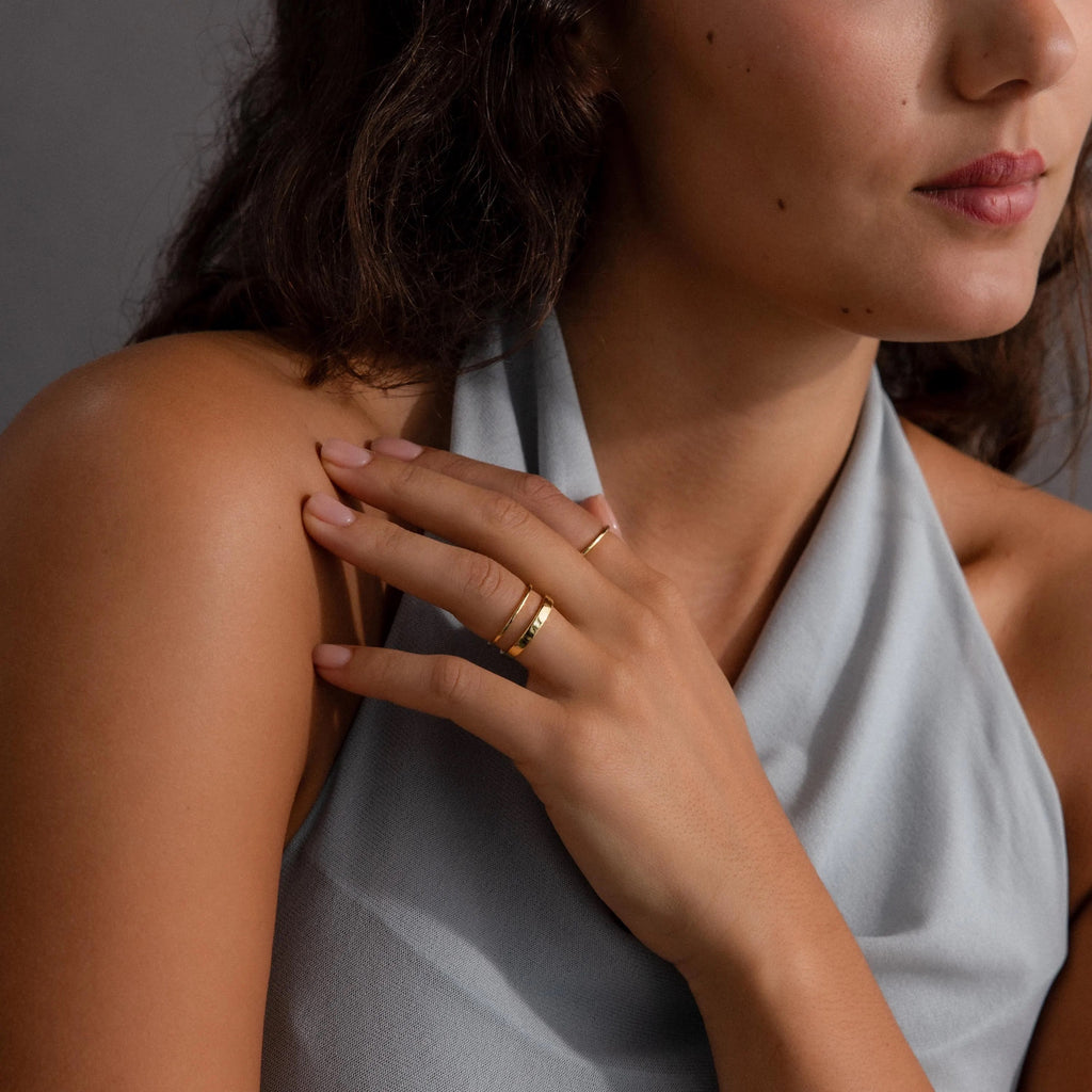 A woman in a gray halter top touches her shoulder, displaying the Simple Ring Set—minimalist gold rings perfect for effortless stacking.